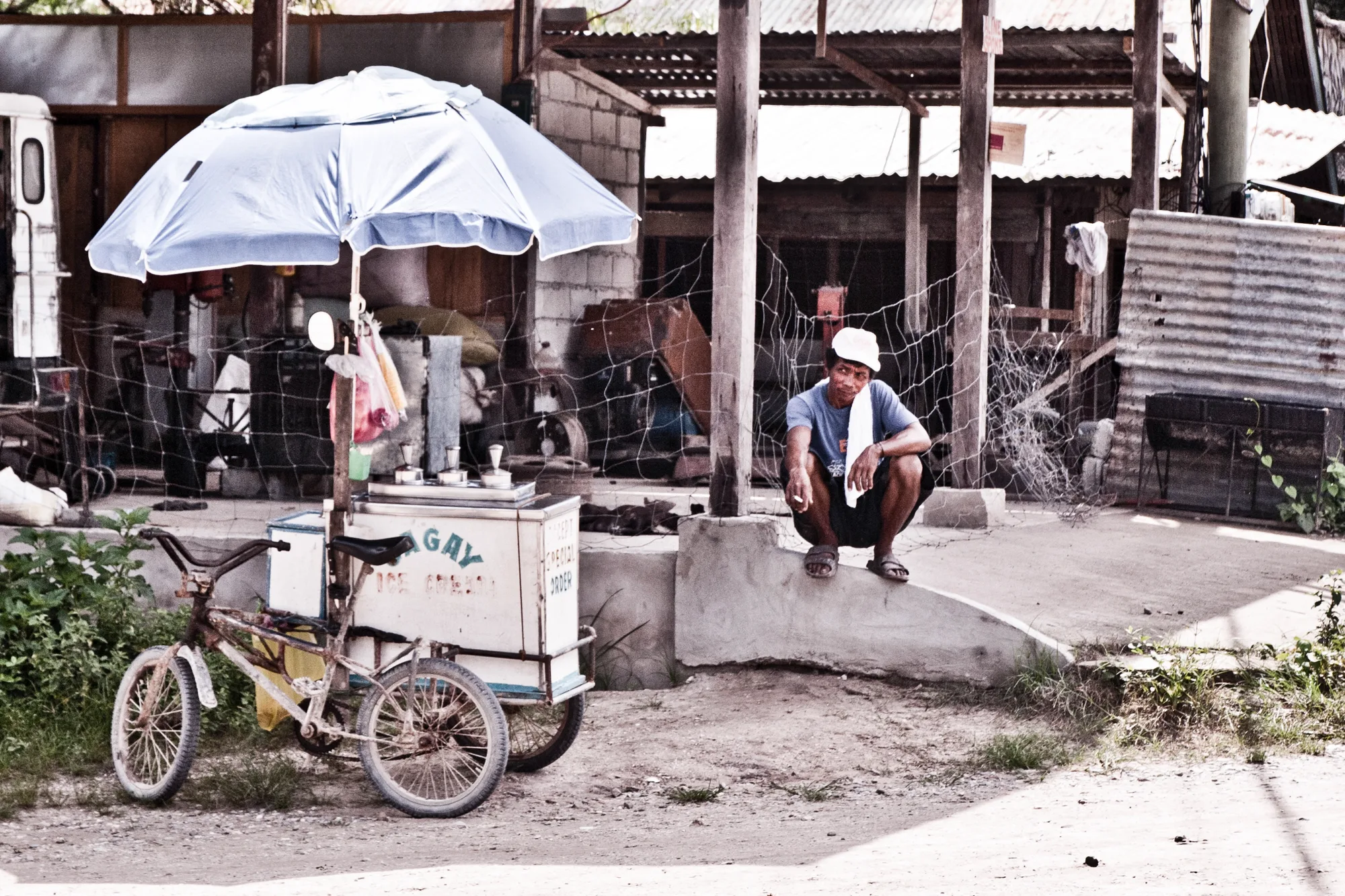 Man squatting beside a bicycle ice-cream cart under a blue umbrella — Palawan, Philippines, documentary and travel photography by Janina Laszlo