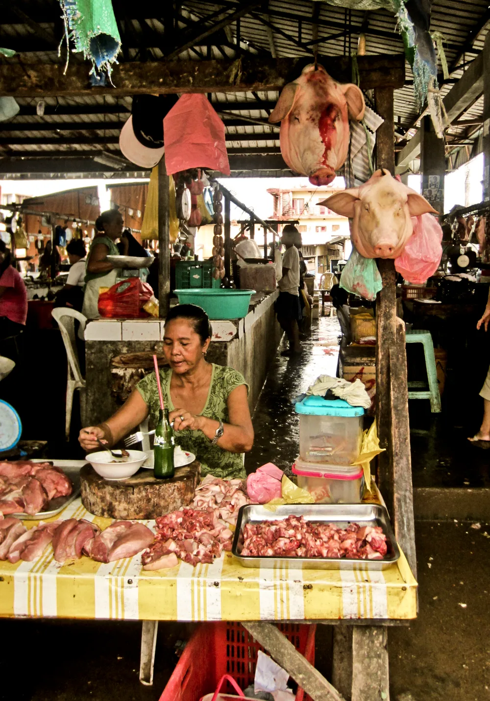 Woman eating at her pork stall below hanging pig heads in a market hall — Palawan, Philippines, documentary and travel photography by Janina Laszlo