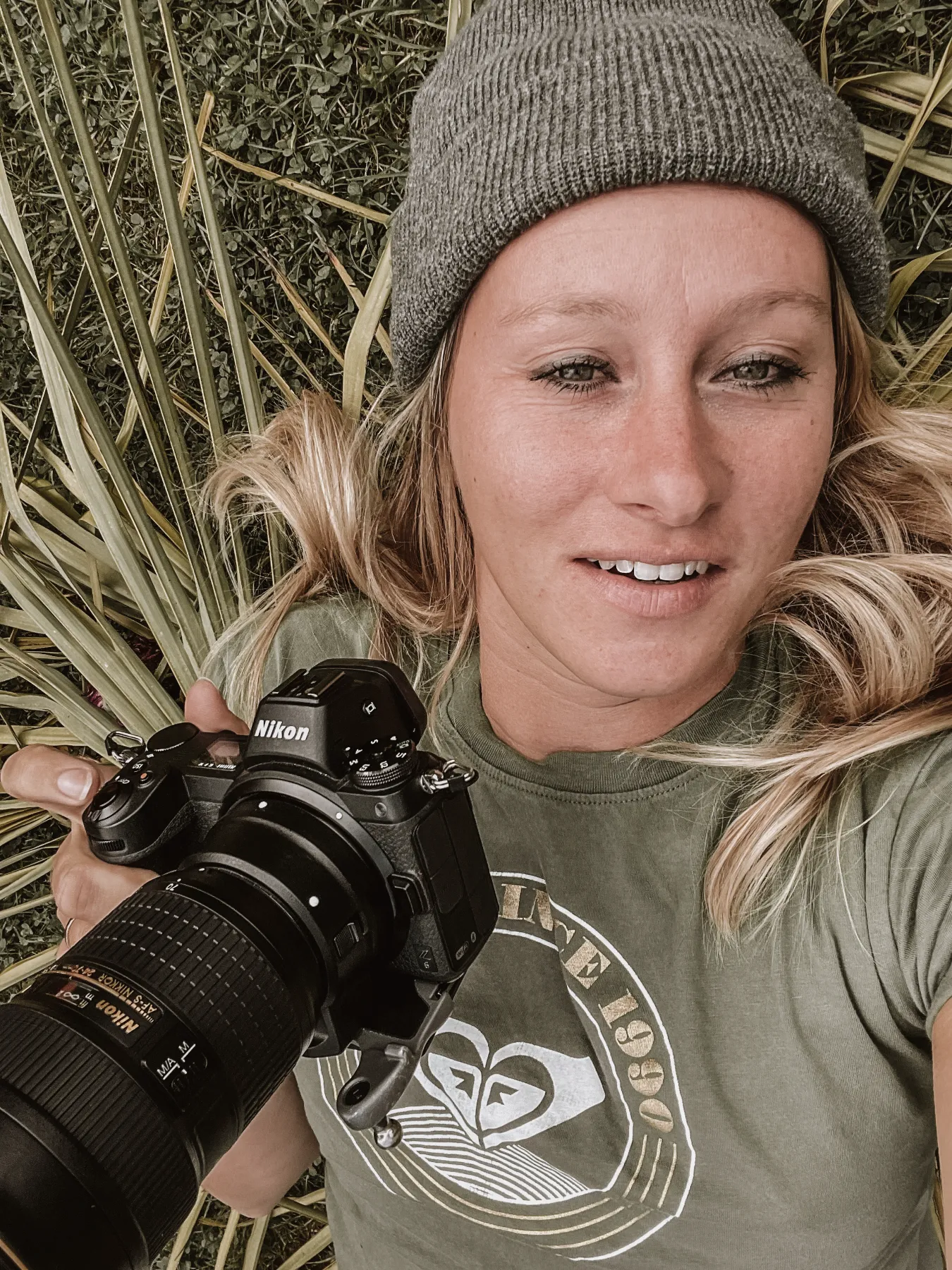 Portrait of Janina Laszlo, photographer in Munich, lying in the grass with her Nikon camera and a grey beanie