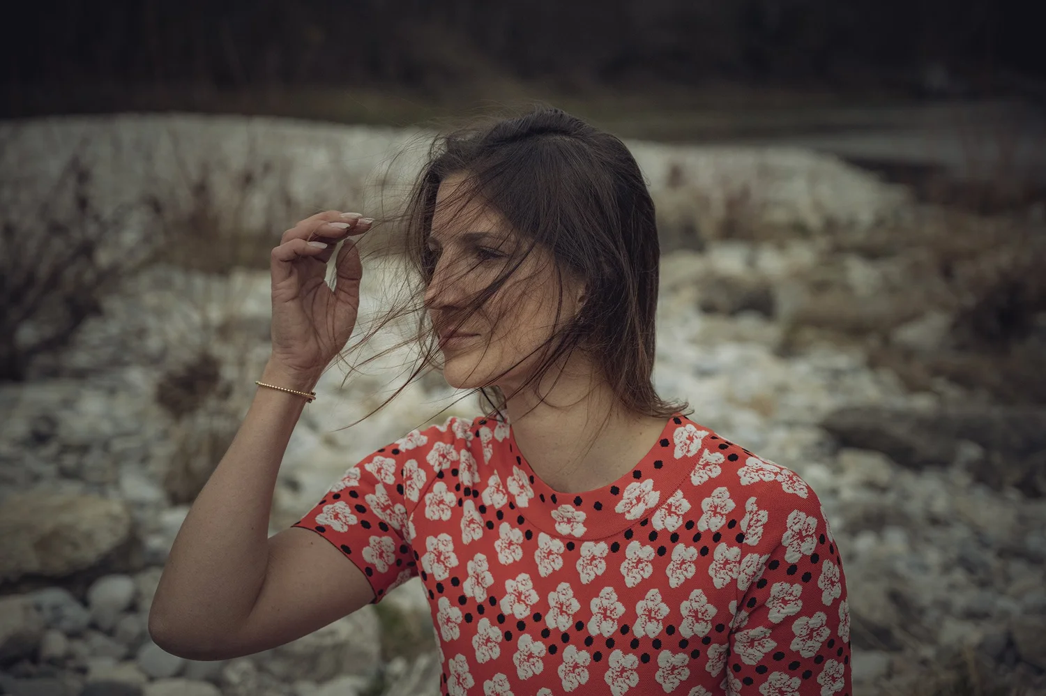 Aline Lettow in a red floral dress brushing hair from her face on a pebble beach — portrait by Janina Laszlo, portrait photographer in Munich