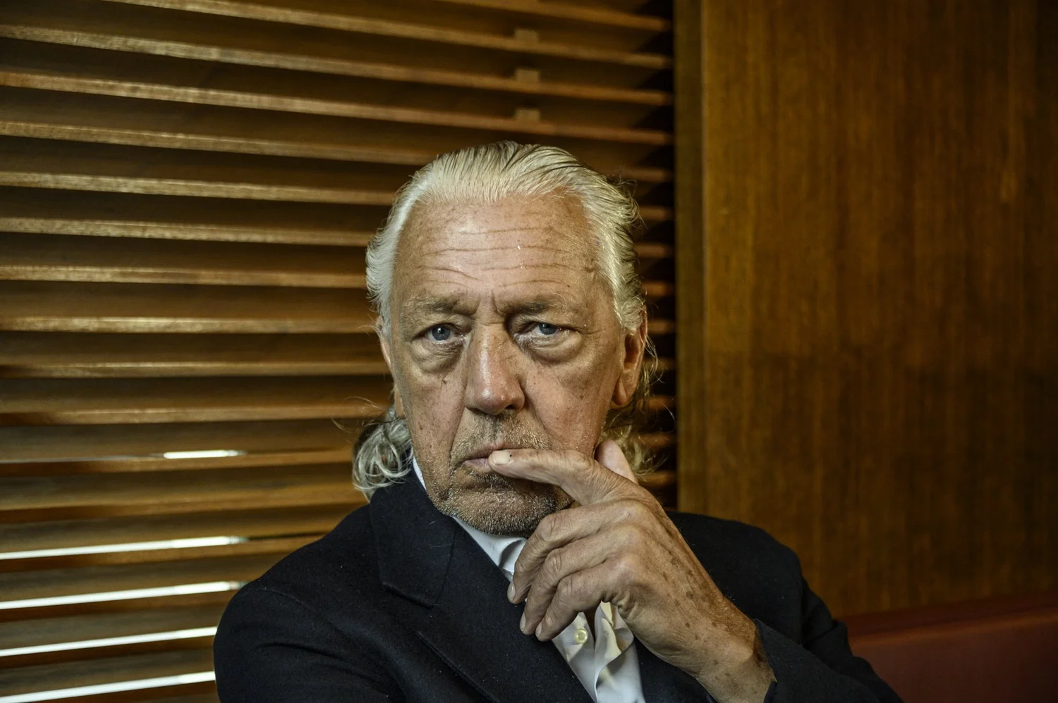 Munich bar legend Charles Schumann in a dark suit in front of wooden blinds — portrait by Janina Laszlo, portrait photographer in Munich