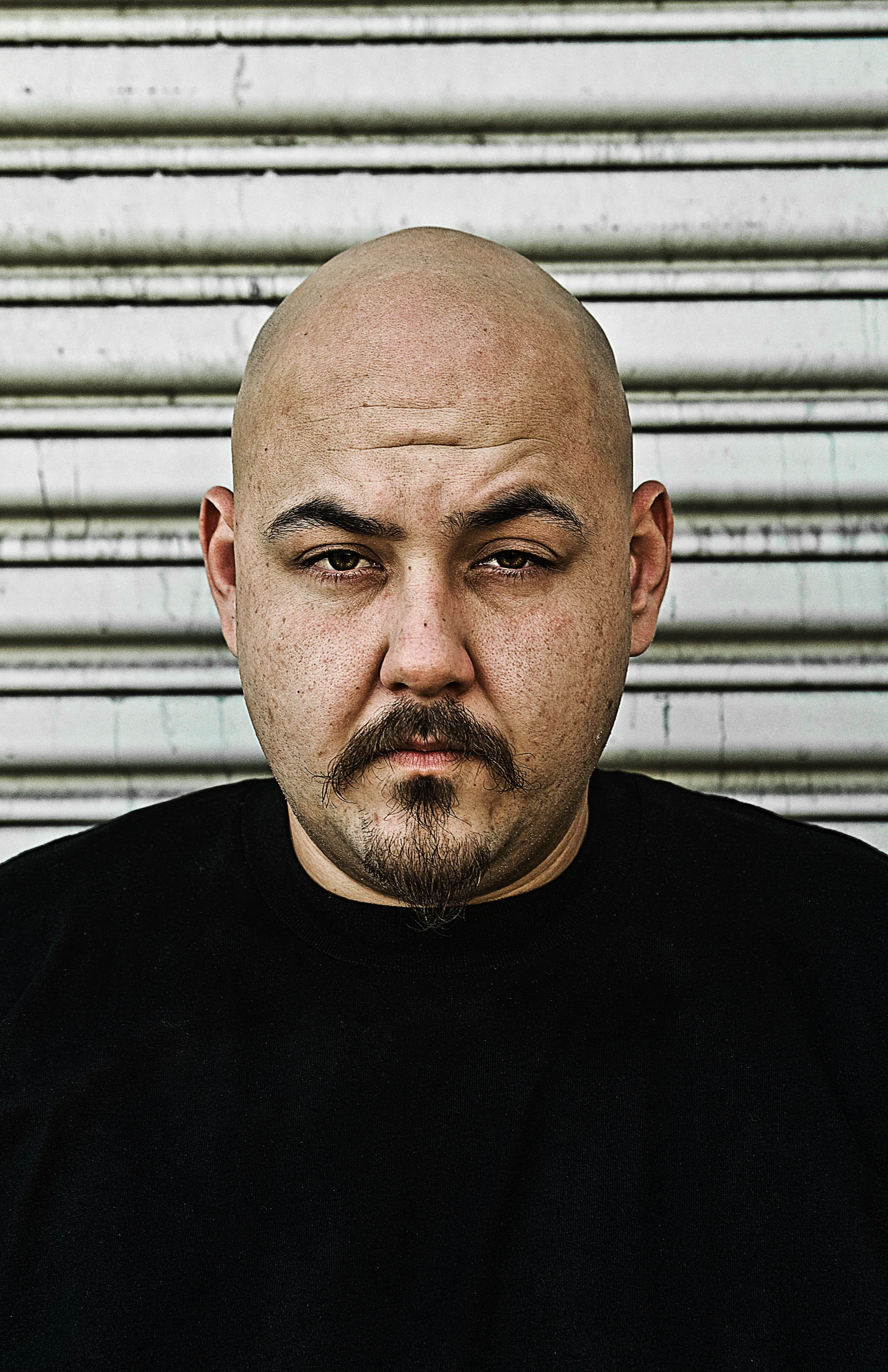 Bald artist in front of a corrugated shutter, direct gaze in East Los Angeles — Suspect Entertainment, music portrait photography by Janina Laszlo