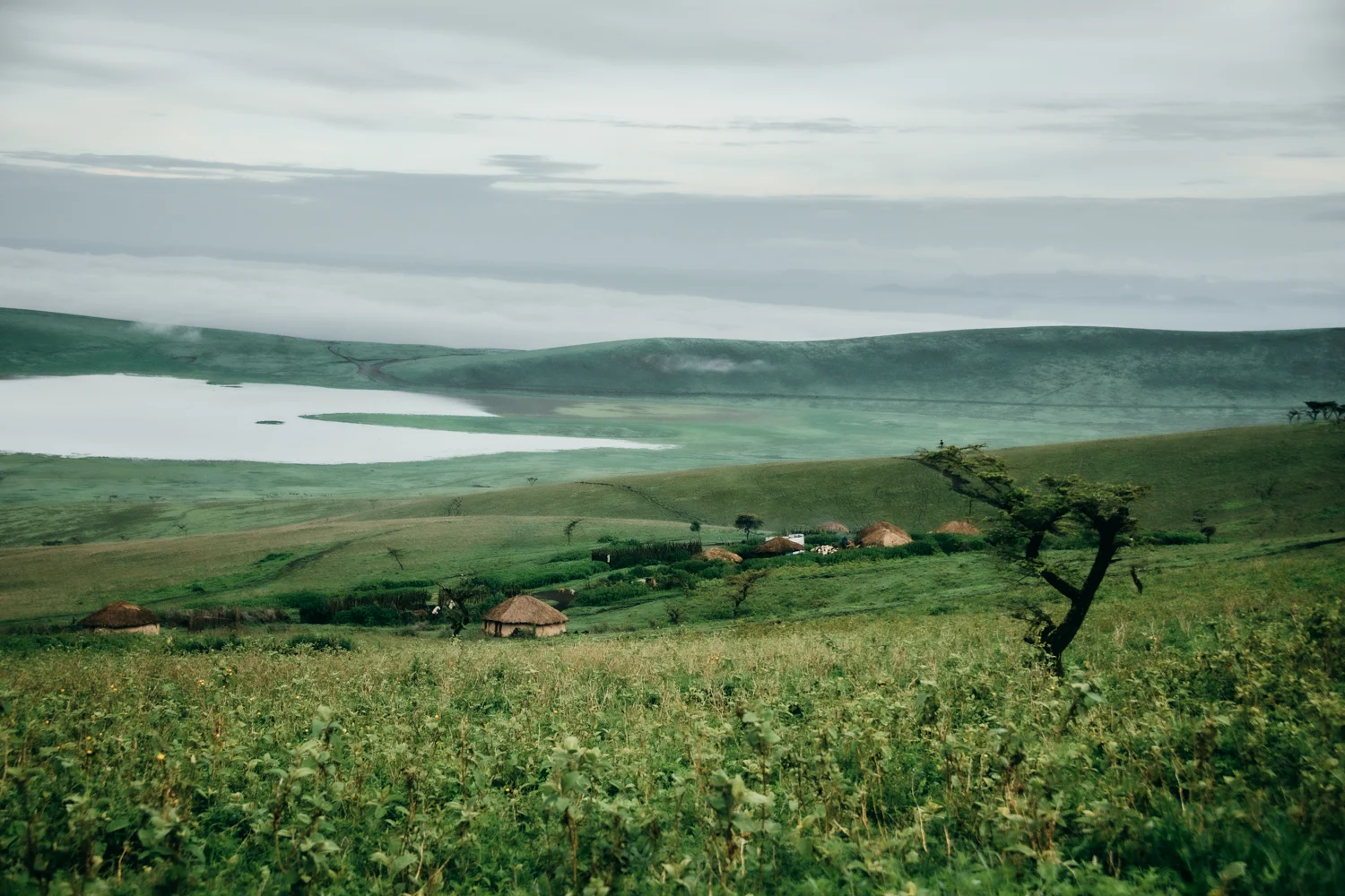 Maasai boma with thatched huts on the crater rim above a lake and green hills, Ngorongoro — Tanzania, travel and wildlife photography by Janina Laszlo