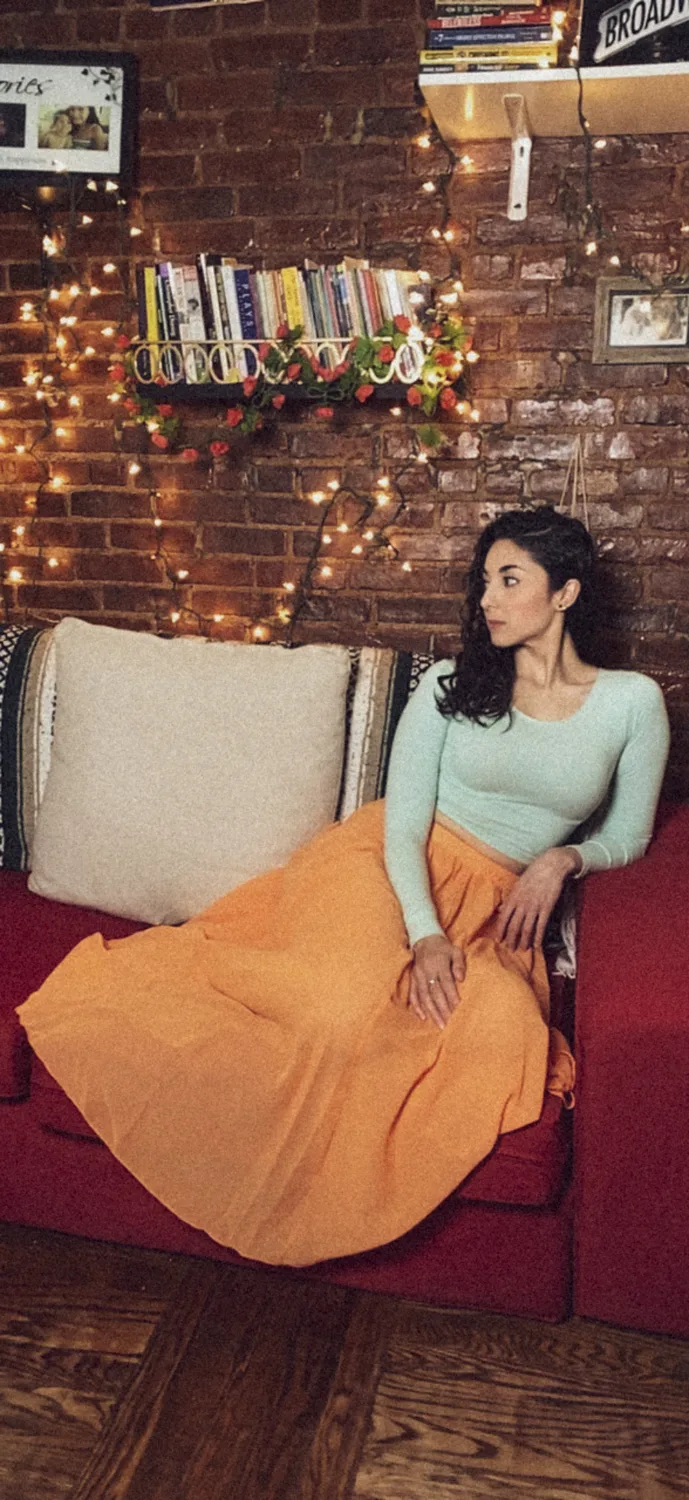 Paula Cossio in an orange skirt on a red sofa against a brick wall with fairy lights, Brooklyn — Deutsche Telekom FaceTime Session, commercial and lifestyle photography by Janina Laszlo