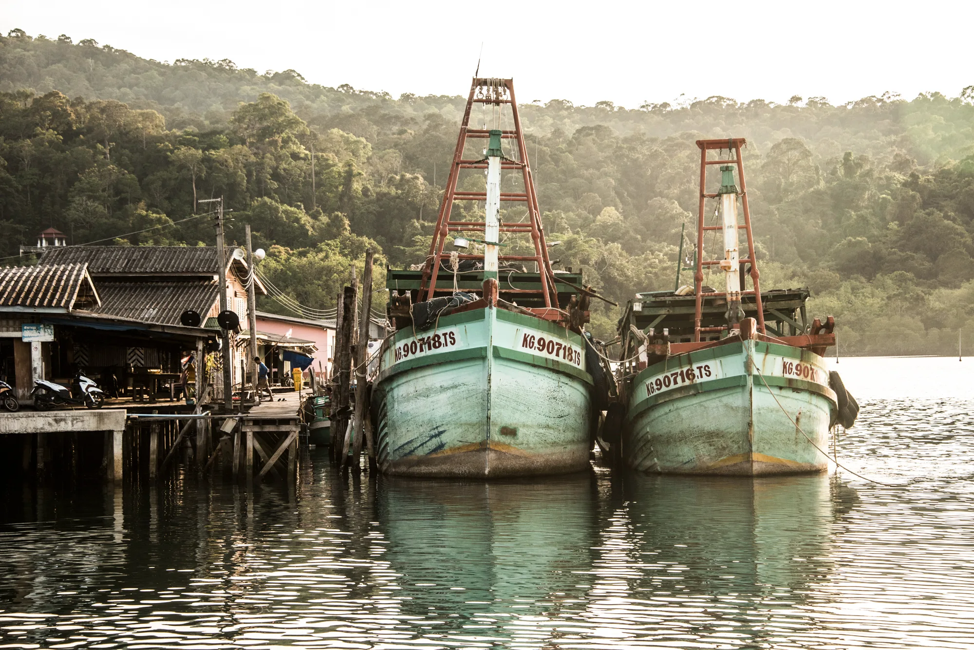Two green trawlers moored beside a stilt village at the foot of jungle hills — Thailand, travel and documentary photography by Janina Laszlo