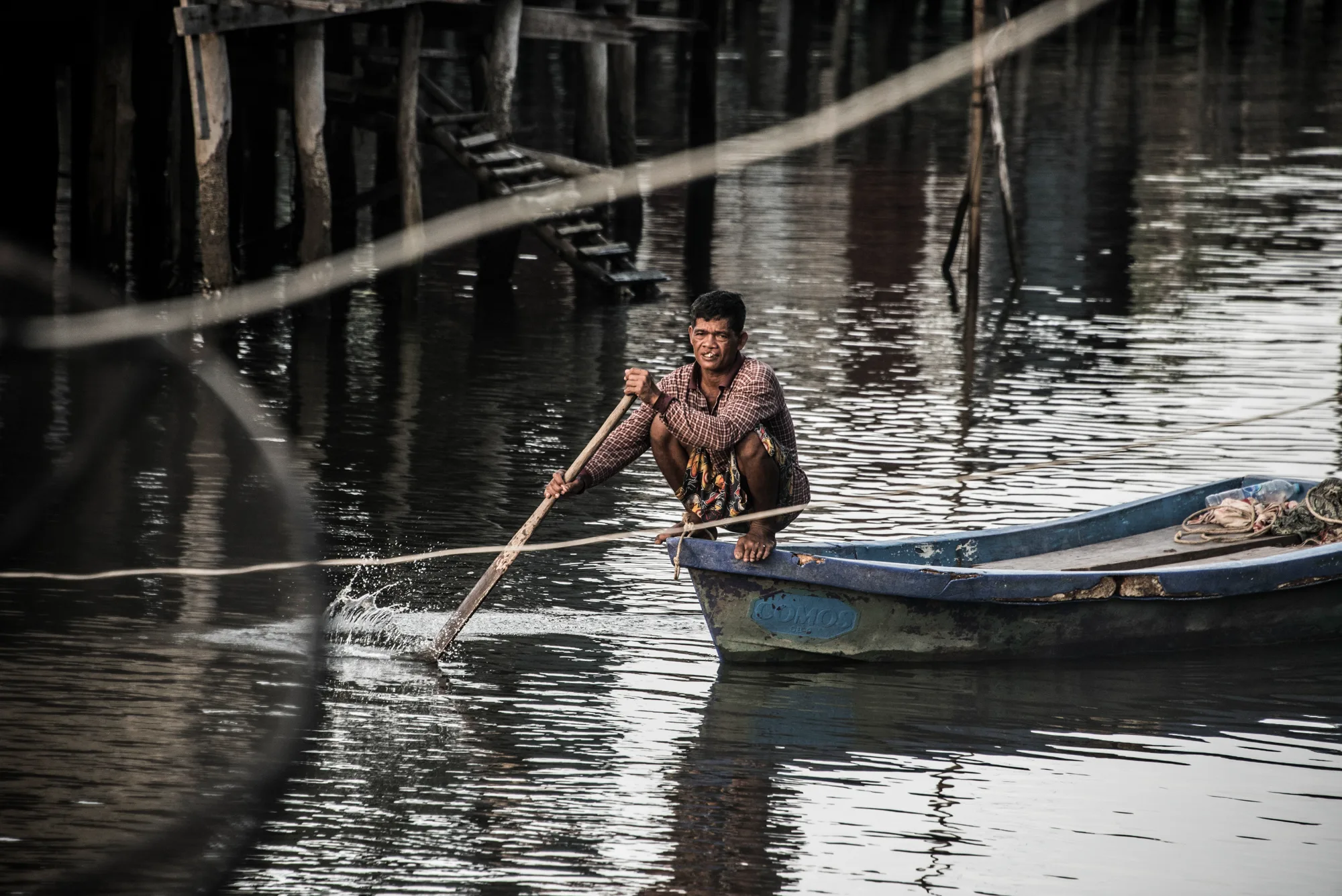 Fisherman squatting on the bow of a blue boat, paddling past stilt houses — Thailand, travel and documentary photography by Janina Laszlo