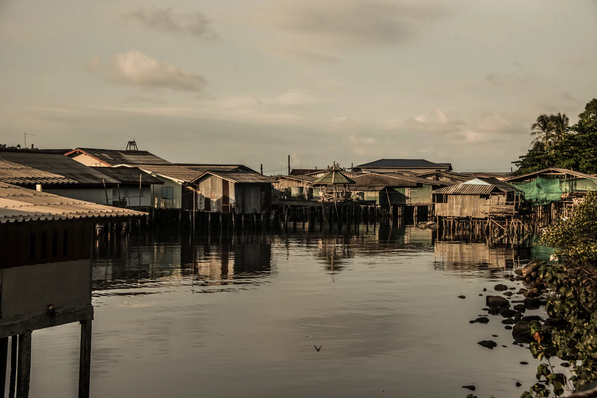 Corrugated-iron stilt houses reflected in a calm inlet at golden hour — Thailand, travel and documentary photography by Janina Laszlo