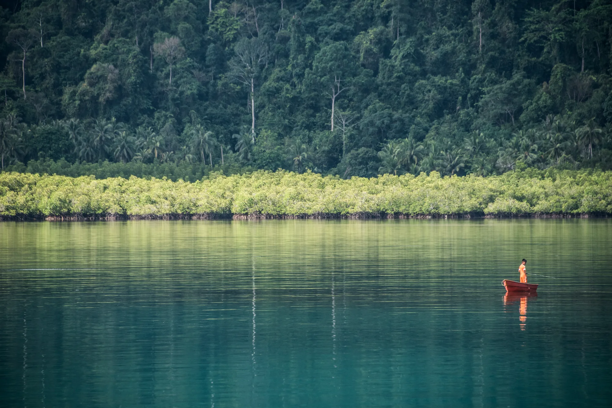 Boy fishing from a red boat in a turquoise bay before mangroves and jungle — Thailand, travel and documentary photography by Janina Laszlo