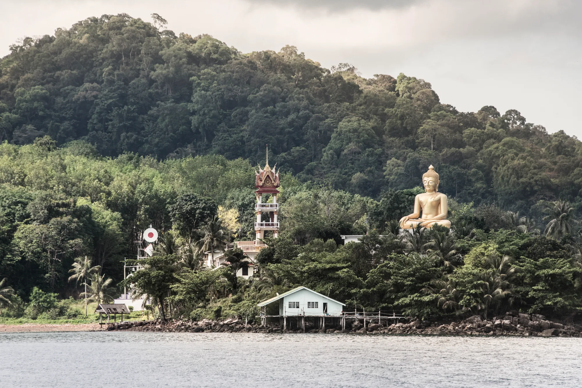 Giant golden Buddha and temple bell tower above a stilt house on the shore — Thailand, travel and documentary photography by Janina Laszlo
