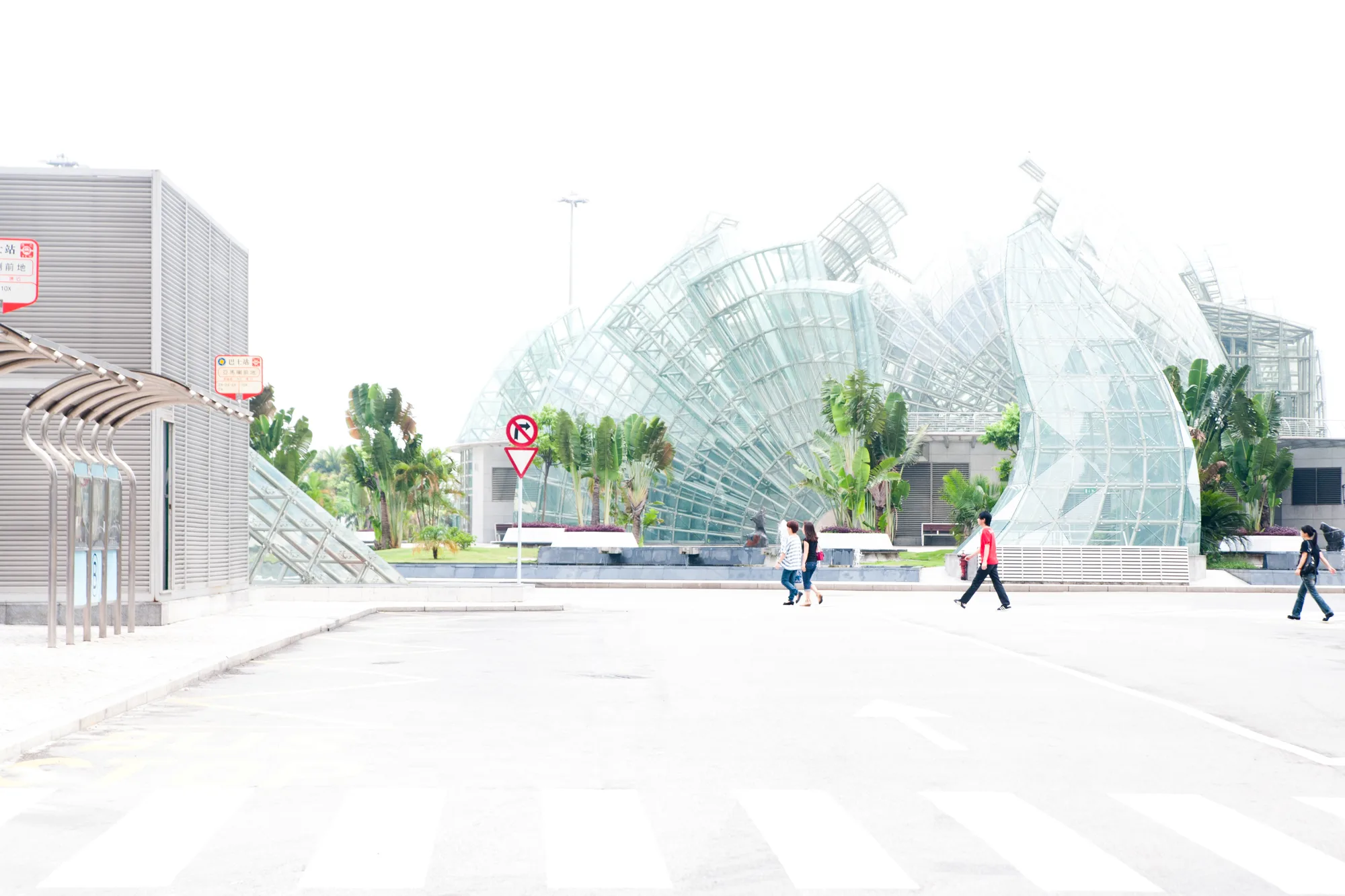 Pedestrians crossing a bright empty plaza past a bus stop and a sweeping glass-and-steel pavilion with palms, Macao — Travel Notes, travel photography by Janina Laszlo