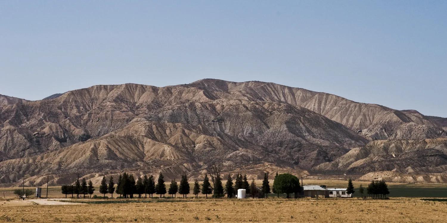 Farmhouse and cypress row before eroded badland mountains, California — US West Coast, panoramic fine art landscape photography by Janina Laszlo