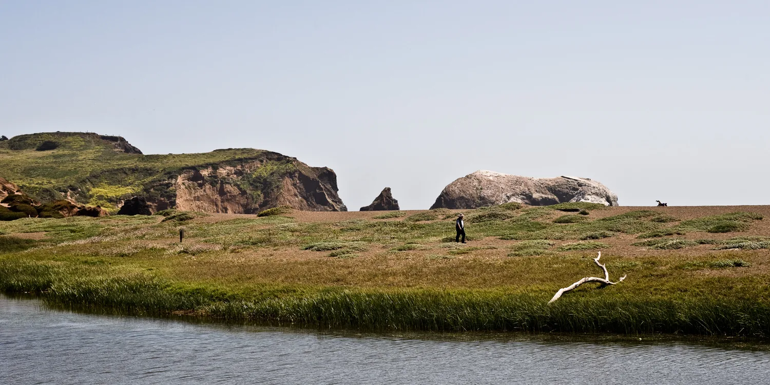 Walker on the grassy shore of Rodeo Lagoon with sea stacks, Marin Headlands, California — US West Coast, panoramic fine art landscape photography by Janina Laszlo