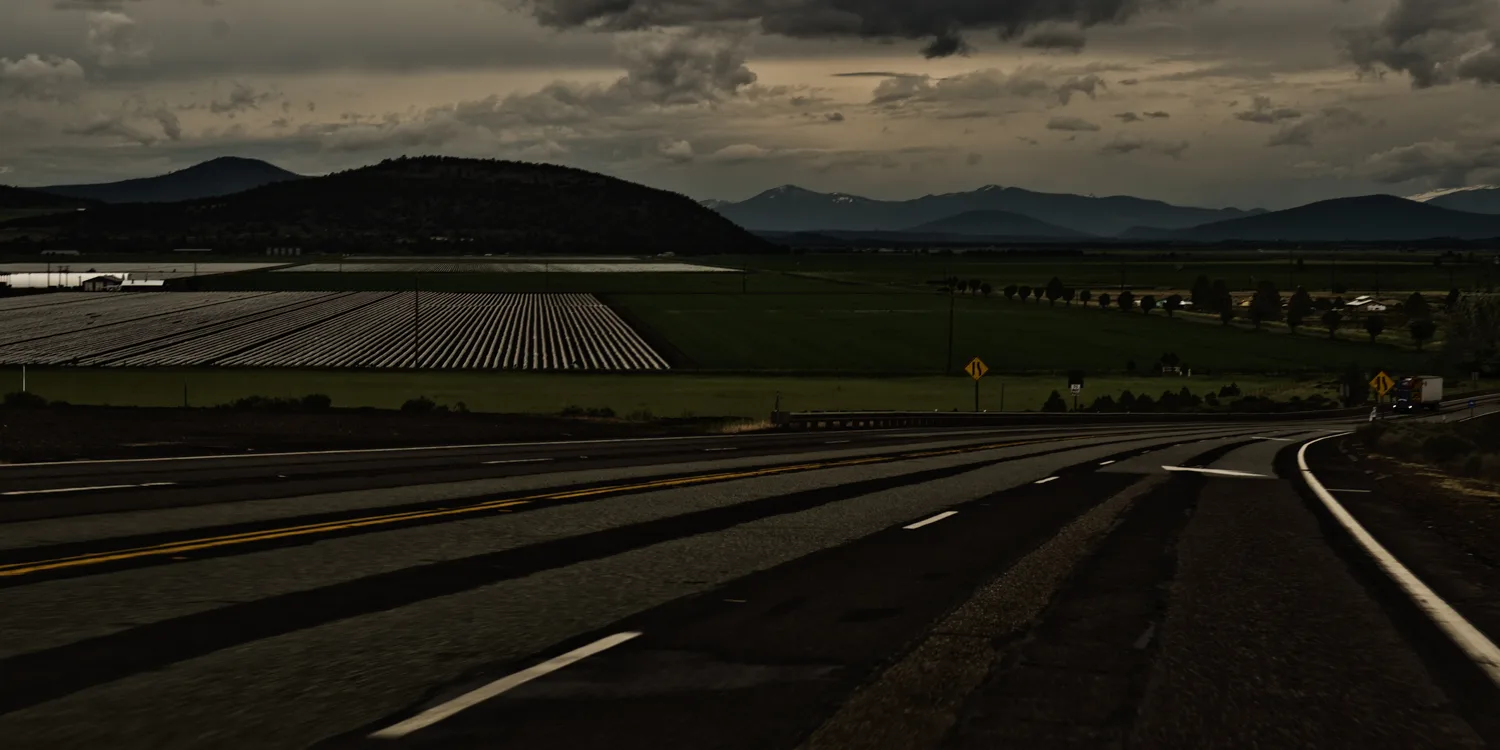 Highway curving past farmland toward the mountains under storm clouds, Northern California — US West Coast, panoramic fine art landscape photography by Janina Laszlo